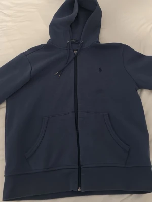 Blå hoodie från Polo Ralph Lauren - Mörkblå hoodie med dragkedja från Polo Ralph Lauren. Klassisk design med huva och dragsko, två fickor framtill och diskret broderad logga på bröstet. Perfekt för en avslappnad och stilren look.
