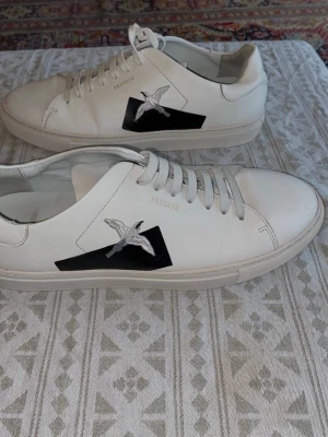 Vita sneakers från Axel Arigato - Snygga vita sneakers från Axel Arigato med svart detalj och broderad fågel på sidan. Skorna har klassisk låg profil, vita snören och är tillverkade i slätt läder. 