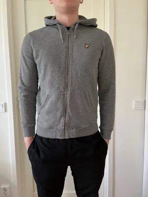 Grå zip hoodie från Lyle & Scott - Snygg grå hoodie med dragkedja från Lyle & Scott. Klassisk passform med huva och dragsko, två fickor fram och den ikoniska gula örnloggan på bröstet. Perfekt för en chill och avslappnad stil. Mjuk bomullsblandning som känns skön mot huden. Den är i hyfsat bra skick men lite använd. Den har två hål på handlederna men annars är den perfekt
