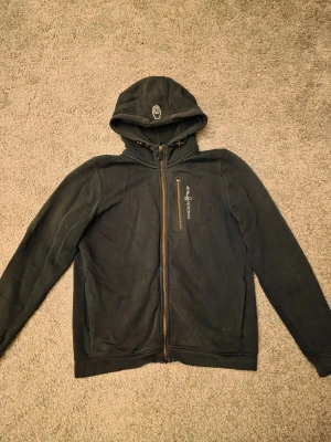 Svart hoodie från Sail Racing med dragkedja - Svart hoodie från Sail Racing med hel dragkedja framtill och huva med tryckt logga. Mjuk fleecekänsla på insidan och en bröstficka med dragkedja. Perfekt för chill dagar eller när du vill ha något bekvämt och stilrent. Dragkedjan på bröstfickan är lite sönder, den man drar i har låstnat därav priset.