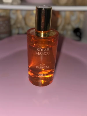 Zara Solar Mango Eau de Parfum - Fräsch parfym från Zara med tropisk känsla. Doften har toppnoter av mango, hjärtnoter av soliga toner, jasmin och kokosnöt samt basnot av sandelträ. Flaskan är orange med guldigt lock och har en modern, rund form.