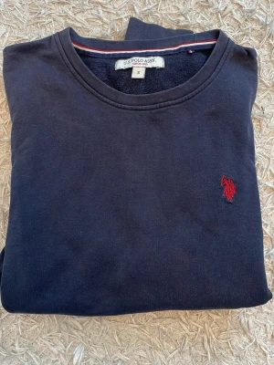 Mörkblå sweatshirt från U.S. Polo Assn - Mörkblå sweatshirt från U.S. Polo Assn med rund halsringning och röd broderad logga på bröstet. Tillverkad i mjukt bomullsmaterial och har ribbade muddar vid ärmslut och nederkant. Enkel och stilren design som passar till det mesta.
