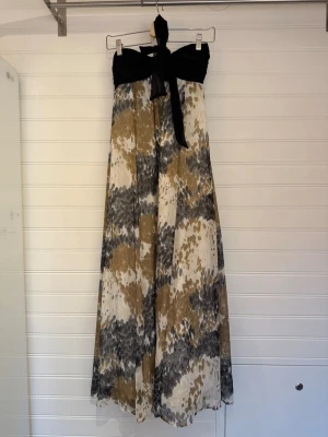Långklänning med svart topp och mönster - Snygg långklänning med svart bandeau-topp och knytband. Kjoldelen har ett abstrakt mönster i beige, grått och vitt. Lätt och flödande material, perfekt för varma dagar. Klänningen är ärmlös och har en feminin siluett.