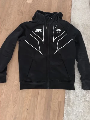Svart UFC hoodie med dragkedja XS - Svart hoodie från UFC med vit logga och grafiska linjer framtill. Dragkedja hela vägen, huva med snören och två fickor. Tillverkad i 55% bomull och 45% polyester. Perfekt för dig som gillar sportig och stilren streetwear.
