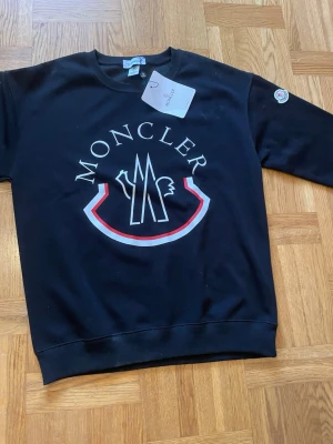 Svart sweatshirt från Moncler - Snygg svart sweatshirt från Moncler med stort vitt och rött logotryck på bröstet och klassisk Moncler-patch på ärmen. Tröjan har rund halsringning, ribbade muddar och är tillverkad i mjukt bomullsmaterial. Perfekt för en trendig och avslappnad stil. Passar storleken S och förmodligen M.