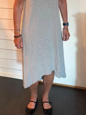 Grå asymmetrisk klänning från COS - Enkel och stilren grå klänning med asymmetrisk nederkant. Klänningen är ärmlös och har en lös passform, perfekt för varma dagar. Tillverkad i mjuk bomull som känns skön mot huden.