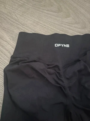 Svarta seamless leggings från DFYNE - Säljer ett par svarta tights från DFYNE med medelhög midja och stretchigt material. Perfekta för gymmet eller löprundan. Tightsen har en diskret logga framtill i midjan och sitter tight för en snygg siluett.