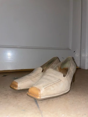 Beige mockaloafers med bruna detaljer - Snygga loafers i beige mocka från Jenny by ara med fyrkantig tå och bruna läderdetaljer framtill. Skorna har låg klack och diskreta sömmar som ger en stilren look. Perfekta för dig som gillar klassisk men trendig stil.