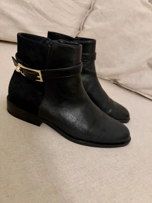 Svarta stövletter med gulddetalj - Snygga svarta stövletter i skinn med dekorativt spänne i guld och pärldetalj vid ankeln. Klassisk modell med låg klack och rundad tå. Perfekta för dig som vill ha en stilren och trendig look.