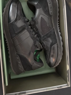 Svarta sneakers från Valentino Garavani - Snygga svarta sneakers från Valentino Garavani med blandning av läder och mocka. Skorna har svarta snören, diskret logga på plösen och coola detaljer på sidorna. Yttersulan är räfflad för extra grepp och stilen är klassiskt sportig men ändå lyxig.