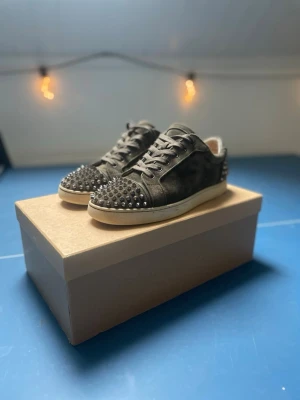 Christian Louboutin  - Säljer ett par mörkgrå sneakers från Christian Louboutin i mocka med coola silverfärgade nitar på tå och häl. Skorna har snörning och vit sula som kontrast. Perfekta för dig som vill sticka ut med en edgy och lyxig stil. hör av dig vid minsta fråga🎉🤩