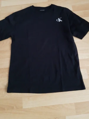 Svart t-shirt från Calvin Klein - En helt ny svart t-shirt från Calvin Klein med klassisk passform och vit CK-logga på bröstet. Enkel och stilren design med rund halsringning och korta ärmar. Perfekt för en clean och modern look. Passar som S är stor i storlek. Passar personer som är 175 till 180. Tveka lnte o fråga om bilder eller kvitto.