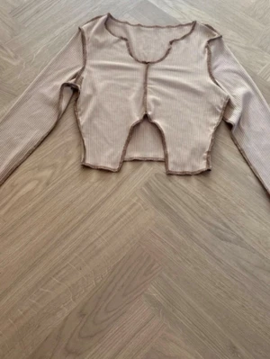 Beige croppad ribbad långärmad topp - Trendig croppad topp i beige med smala ränder och råa kanter. Toppen har lång ärm, ribbad struktur och en unik skärning framtill. Perfekt för att skapa en cool och avslappnad look.