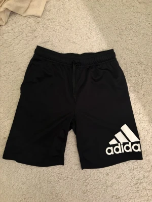 Svarta Adidas shorts med logga - Säljer ett par svarta shorts från Adidas med stor vit logga på vänster ben. Shortsen har elastisk midja med snörning och är tillverkade i ett mjukt och lätt syntetmaterial, perfekta för träning eller chill.