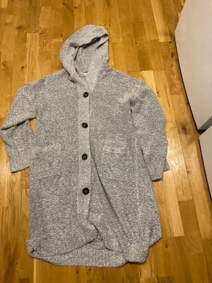Grå lång kofta med huva och knappar stl XSmall  - Mysig lång kofta i gråmelerat material med stora svarta knappar framtill. Koftan har huva, två framfickor och långa ärmar. Perfekt för chill hemma och loungewear. Materialet ser ut att vara mjukt och stickat.