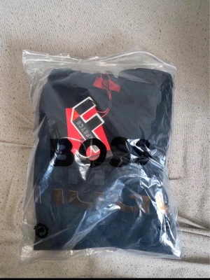 Svart Hugo Boss tshirt dress - Snygg svart hoodie från Hugo Boss med stort HUGO-tryck i guld och svart framtill. Klassisk modell med huva och ribbade muddar. Perfekt för dig som vill ha en stilren och trendig look.