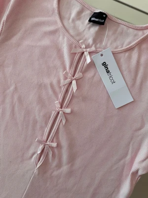 Ljusrosa ribbad topp med rosetter - Supersöt ljusrosa ribbad topp från Gina Tricot med dekorativa rosetter framtill. Toppen har rund halsringning och trekvartsärm, perfekt för en trendig och feminin look.