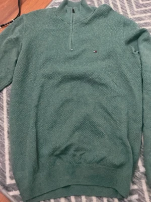 Grön halvzip tröja Tommy Hilfiger - Snygg grön stickad tröja från Tommy Hilfiger med halvzip och diskret logga på bröstet. Tröjan har en strukturerad yta och ribbade muddar vid ärmslut och nederkant. Perfekt för dig som gillar klassisk men avslappnad stil.PRIS KAN DISKUTERAS