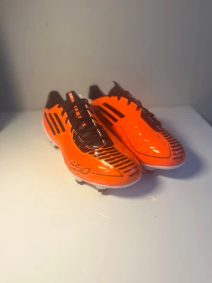 Adidas F50 orange fotbollsskor FG (äldre modell) - Snygga Adidas F50 fotbollsskor i en skarp orange färg med svarta detaljer och vita sulor. Skorna har klassiska svarta ränder på sidan, snörning och dobbar för bra grepp på planen. Tillverkade i syntetmaterial för lätt känsla och snabbhet.