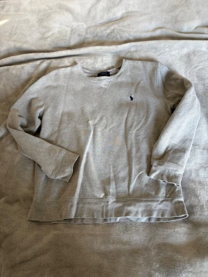 Grå sweatshirt från Polo Ralph Lauren - Klassisk grå sweatshirt från Polo Ralph Lauren med broderad logga på bröstet. Tröjan har rund halsringning, långa ärmar och ribbade muddar. Perfekt för en avslappnad och stilren look.