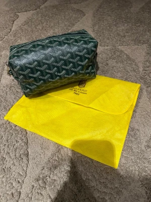 Grön handväska med mönster - Snygg grön handväska med  ikoniska vita mönster och logga. Väskan har en mjuk, rektangulär form och är tillverkad i canvas med detaljer i läder. Kommer med en gul dustbag med Goyard Paris-tryck.
