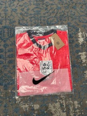 Röd England fotbollströja Nike - Säljer en röd England fotbollströja från Nike med svart och vit detalj vid krage och ärmslut. Tröjan har England-emblemet på bröstet och ett svart Nike-märke. Tillverkad i lätt och ventilerande material, perfekt för träning eller match.