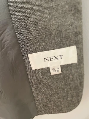 Grå kavaj från Next, storlek 36 - Stilren grå kavaj från Next i storlek 36. Klassisk modell med smal passform, helfodrad insida och knappar vid ärmsluten. Materialet är en mjuk och lätt vävd textil som ger en snygg och modern look.