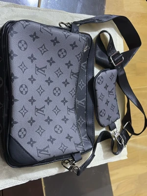  axelväska svart - Snygg axelväska i svart och grått med klassiskt monogrammönster. Väskan har justerbar axelrem i textil och en mindre avtagbar ficka. Tillverkad i tåligt lädermaterial med silverfärgade detaljer och dragkedja.