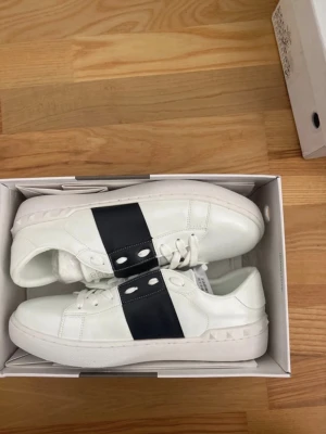 Valentino vita sneakers med svart band - Snygga vita sneakers från Valentino med ett brett svart band över vristen och guldfärgad logga på plösen. Skorna har vita snören, rund tå och detaljerade nitar längs sulan. Tillverkade i slätt läder för en clean och modern look.