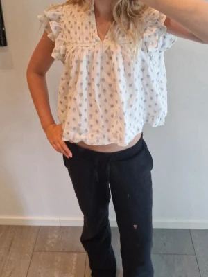 Vit blus med volanger och blått mönster - Söt vit blus med små blå blommönster och volangdetaljer vid axlarna. Blusen har v-ringning och korta ärmar med rysch. Luftig och lätt i materialet, perfekt för sommardagar. Passar till både jeans och kjol för en avslappnad look. Aldrig andvänd även om lappen är borta.