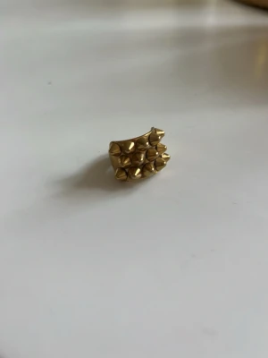 Guldfärgad edblad ring med nitar - Ring i guldfärgad metall med tre rader av spetsiga nitar framtill. Ringen har en chunky och edgy design som verkligen sticker ut och ger en tuff känsla till din stil. Perfekt för dig som gillar statement-smycken och vill ha något unikt.