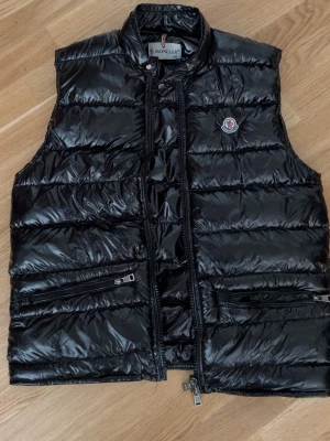 Svart dunväst från Moncler - Snygg svart dunväst från Moncler med glansig finish och klassisk logotyp på bröstet. Västen har hög krage, två dragkedjeförsedda fickor framtill och stängs med dragkedja. Perfekt för lager på lager och streetwear-stil.