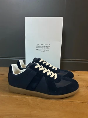 Maison Margiela navy sneakers - Snygga sneakers från Maison Margiela i mörkblå mocka och läder med vita skosnören och beige gummisula. Klassisk low-top siluett som passar till många olika stilar. Diskret branding och stilren design.