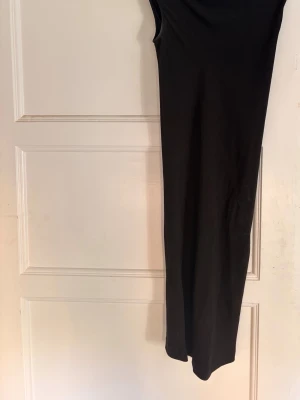 Svart långklänning med dragkedja - Stilren svart långklänning från Rick Owens med en markerad dragkedja bak som ger en edgy touch. Klänningen har en rak passform med ett mer flowigt, hängande del rund bröstet. Klänningen är tillverkad i ett mjukt material som sitter snyggt på kroppen. Perfekt för dig som gillar minimalistisk och modern stil. 