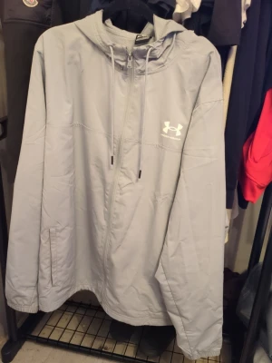 Ljusgrå vindjacka från Under Armour - Ljusgrå vindjacka från Under Armour med huva och dragkedja framtill. Jackan har två sidofickor, justerbar dragsko i huvan och en stilren logga på bröstet. Perfekt för blåsiga dagar och sportiga outfits.
