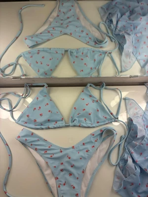 Ljusblå bikiniset  med små blommor och sjal i samma tyg - Supersöt ljusblå bikiniset med små röda/rosa blommor och tunna knytband. Klassisk trekantsmodell upptill och högt skurna trosor. Materialet känns mjukt och stretchigt.        Perfekt för stranden eller poolen. Endast använd vid ett tillfälle!☀️