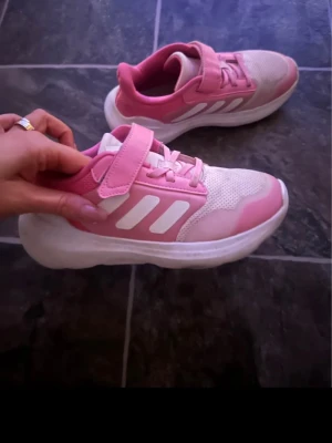 Rosa och vita sneakers från Adidas storlek 33 - Snygga sneakers från Adidas i rosa och vitt med klassiska tre ränder på sidan. Skorna har både kardborreband och snörning för extra bra passform. Ovandelen är i mesh och syntetmaterial, vilket ger bra andningsförmåga och komfort. Perfekta för en sportig och trendig look.