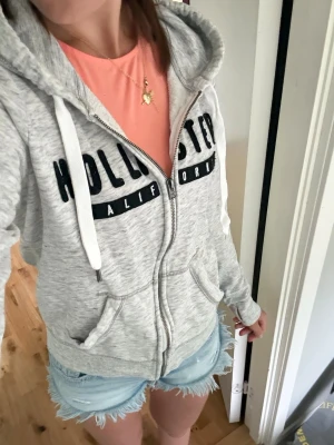 Grå zip hoodie från Hollister - Snygg ljusgrå hoodie från Hollister med dragkedja framtill och stor svart logga över bröstet. Hoodien har vita snören i huvan, två fickor fram och ribbade muddar vid ärmar och nederkant. Perfekt för en avslappnad och trendig look.