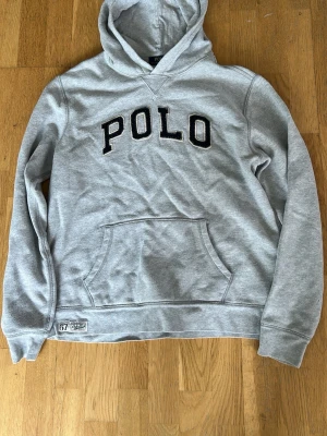 Grå hoodie från Polo Ralph Lauren - Snygg grå hoodie från Polo Ralph Lauren med stor svart POLO-text broderad på bröstet. Klassisk känguruficka framtill och huva. Tillverkad i mjukt bomullsmaterial som känns skönt mot huden. Perfekt för en avslappnad och sportig stil.