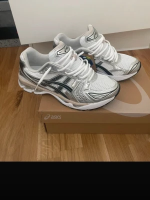Asics Gel Kayano 14 - Snygga vita och silverfärgade sneakers från ASICS med mesh- och syntetmaterial. Skorna har klassisk låg profil, vita snören och svarta detaljer på sidan. Perfekta för dig som gillar sportig och trendig stil.