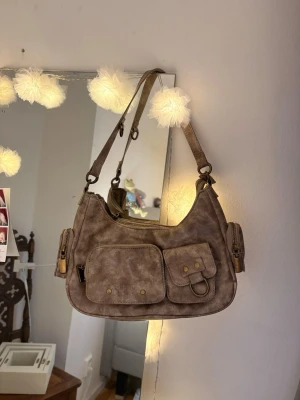 Beige/brun axelremsväska/handväska med fickor - Snygg beige handväska i fejkskinn med flera praktiska ytterfickor och guldfärgade detaljer. Går perfekt att få plats med AirPods på sidorna!🥰 Väskan har justerbar axelrem och dragkedja upptill. Perfekt för dig som gillar en cool och funktionell stil med plats för det viktigaste 