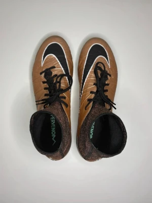 Nike Hypervenom Phantom 2 - Snygga fotbollsskor från Nike i brons med svart och vitt mönster. Skorna har högt skaft i stickat material för extra stöd runt ankeln, svarta skosnören och dobbar för bra grepp på planen. Perfekta för dig som vill sticka ut på fotbollsplanen.