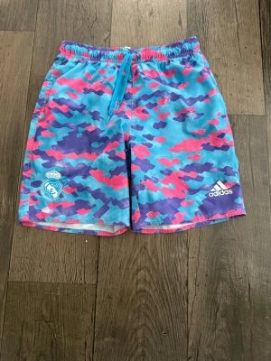Färgglada Real Madrid badbyxor Adidas - Säljer ett par färgglada badbyxor från Adidas x Real Madrid med blå, lila och rosa camomönster. De har resår och dragsko i midjan samt innerbyxa i mesh. Tillverkade i Primegreen-material för en mer hållbar känsla. Perfekta för stranden eller poolen!