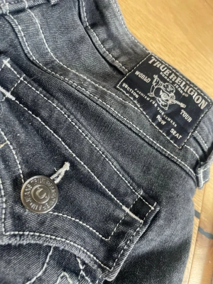 Svarta jeansshorts True Religion - Säljer ett par svarta jeansshorts från True Religion med slitna kanter och kontrastsömmar. Klassisk femficksmodell med knapp och dragkedja framtill. Snyggt tvättade detaljer och logga på bakfickan. Perfekta för sommaren!