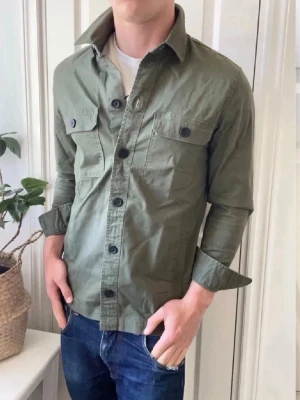 Hollister overshirt - En overshirt från Hollister med knäppning framtill och två bröstfickor med lock och knappar. Storlek S. Långärmad modell i lätt bomullstyg, perfekt att bära öppen eller stängd över en t-shirt. Enkel och stilren design som passar till jeans.