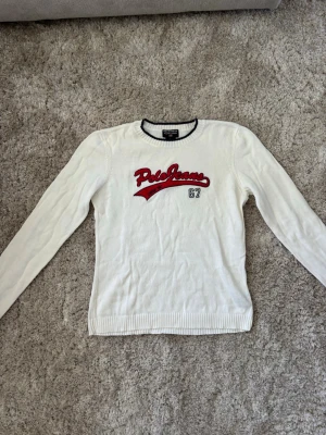 Vit stickad tröja Polo Ralp Lauren Jeans  - Vit stickad tröja från Polo Jeans Co. med röd broderad logga och texten '67' på bröstet. Tröjan har rund halsringning med mörkblå kant och långa ärmar med ribbade muddar. Perfekt för en avslappnad och sportig stil.