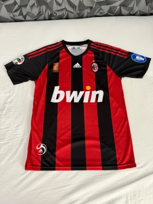 AC Milan Ronaldinho matchtröja - Säljer en klassisk AC Milan matchtröja från Adidas med Ronaldinho och nummer 80 på ryggen. Tröjan har röda och svarta vertikala ränder, vita tryck och flera officiella klubbmärken och sponsorer. Perfekt för dig som älskar fotboll och vill ha en ikonisk supportertröja.
