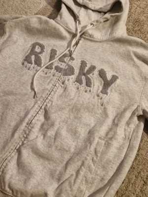 Grå hoodie med dragkedja och text - Säljer en ljusgrå hoodie med dragkedja från Risky. På bröstet står det 'RISKY' i stora, fluffiga bokstäver med drip-effekt. Hoodien har huva med dragsko och fickor framtill. Materialet är mjukt och skönt, perfekt för chill dagar. Storlek Small herr, kontakta mig för mer info!