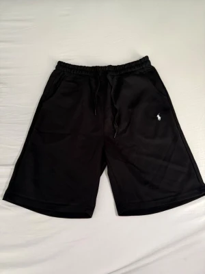 Svarta shorts från Polo Ralph Lauren - Svarta shorts från Polo Ralph Lauren med klassisk logga broderad i vitt på vänster ben. Modellen har elastisk midja med snörning och två sidofickor. Tillverkade i mjuk bomull, perfekta för en avslappnad stil. Helt nya med ettiket.