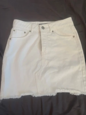 Vit jeanskjol med fransig kant - Säljer en vit kort jeanskjol med rå, fransig kant nertill och klassiska fickor fram. Kjolen har knapp och dragkedja i midjan och är perfekt för en avslappnad och trendig look.